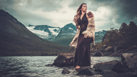 A majestic woman dressed in a flowing gown and fur-trimmed cloak stands barefoot on the rocky edge of a lake. Snow-capped mountains rise in the background under a cloudy sky, evoking themes of sovereignty and connection to nature.